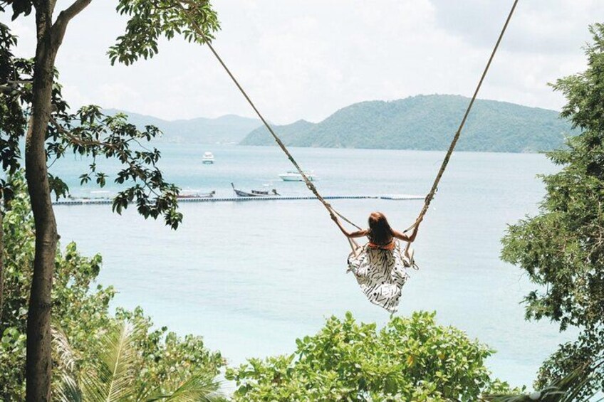 A person swinging on a rope swing over a body of water with boats and mountains in the background. 2