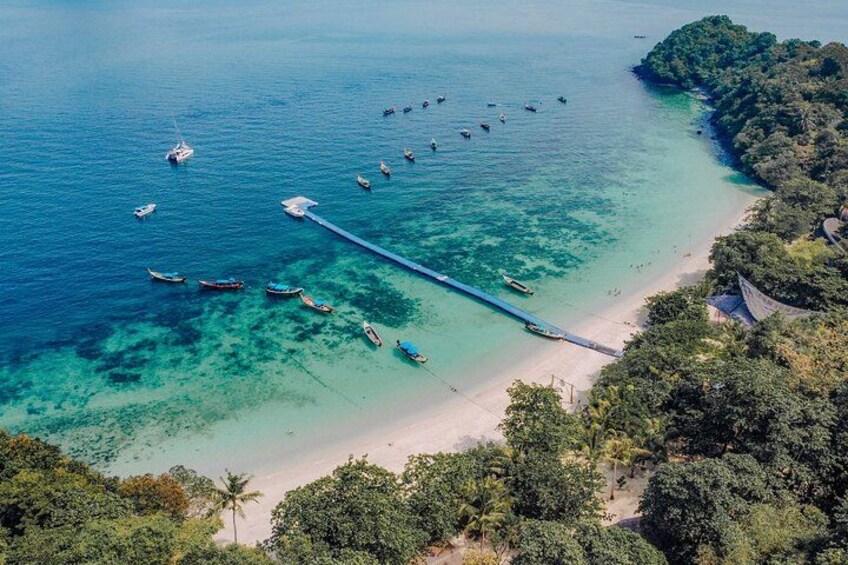 A coastal landscape with a clear turquoise sea, a sandy beach, and a long pier extending into the water. 4