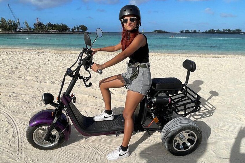 A person riding a three-wheeled electric scooter on a sandy beach with the ocean and a distant island in the background. 1