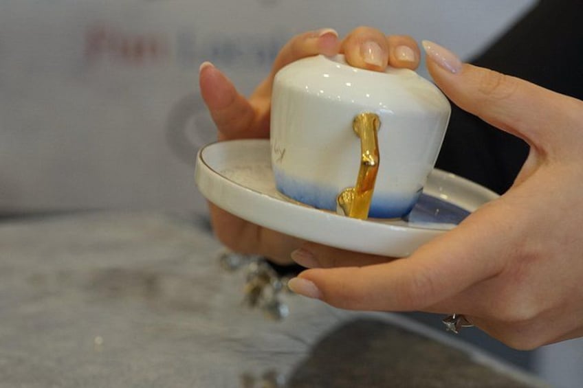 A person holding a white teacup with a gold handle, placed on a saucer. 3