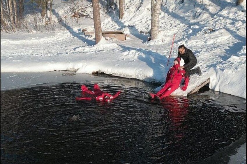 En person i en röd snöoverall står på en frusen sjö, en annan person i en svart jacka hjälper till, och en röd pulka står i närheten. 3