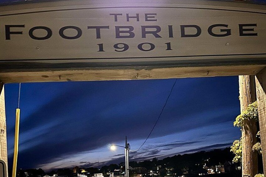 A street sign reading 'The Footbridge 1901' with a night sky and city lights in the background. 1