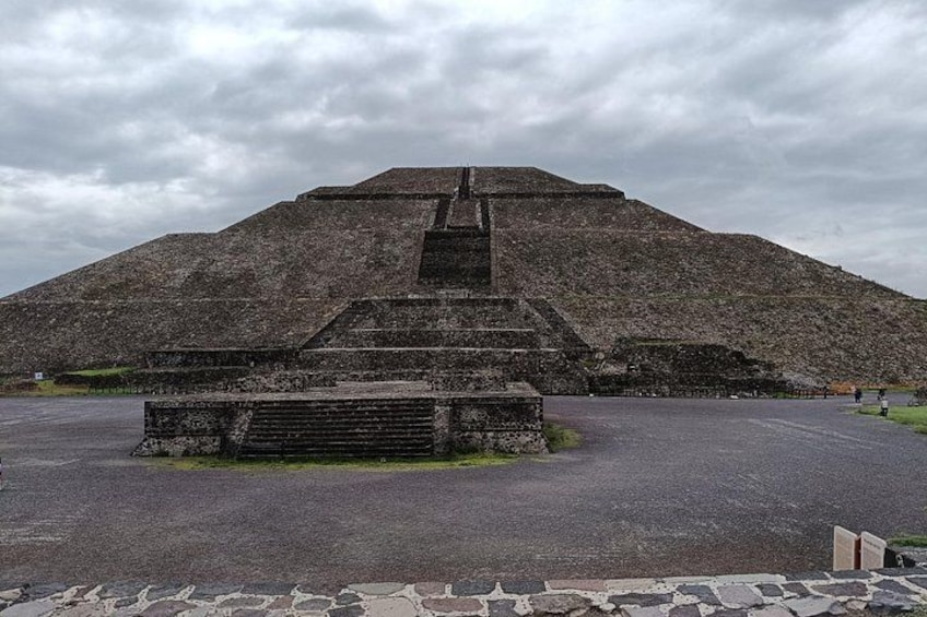 Eine große pyramidenförmige Struktur mit einer Treppe, die zum Gipfel führt, umgeben von einer gepflasterten Fläche. 1