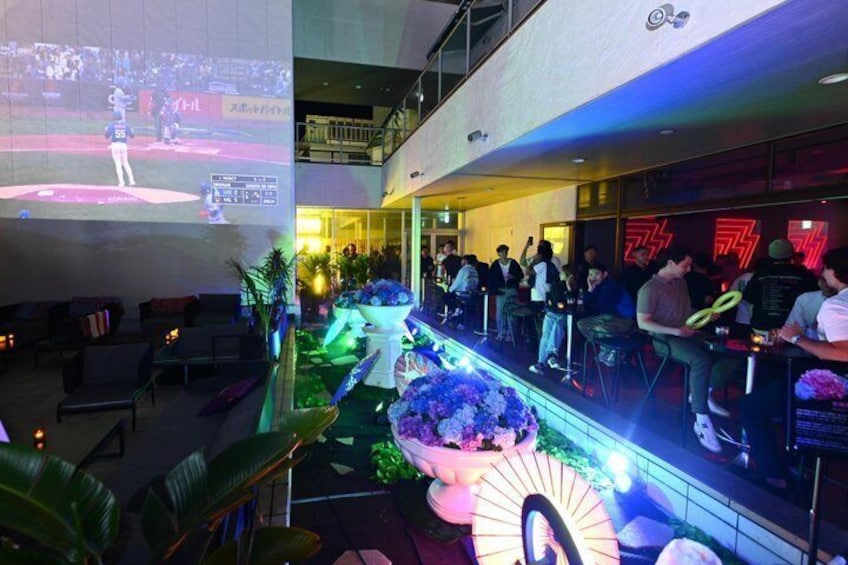 Een sportbar met een groot scherm waarop een honkbalwedstrijd wordt vertoond, een bargedeelte met neonborden en een zithoek met planten. 1