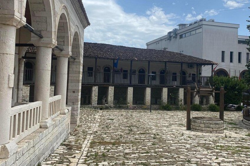 A courtyard with stone paving, a building with arches, and a covered area with a tiled roof. 4