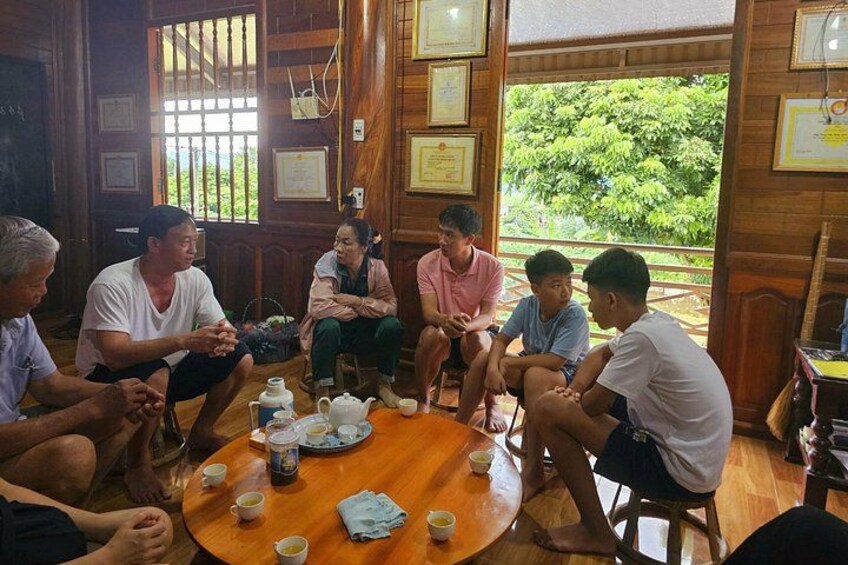 A group of people sitting around a wooden table in a room with large windows, some holding cups. 1
