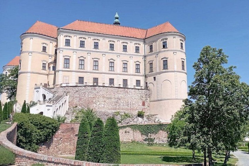 A large, historic building with a red roof, surrounded by well-maintained gardens and a stone wall. 2