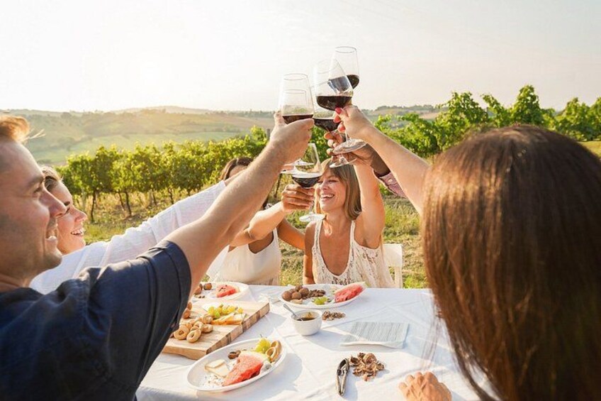 Un groupe de personnes porte un toast avec des verres de vin autour d’une table où se trouve de la nourriture, en plein air, avec un vignoble en arrière-plan. 3