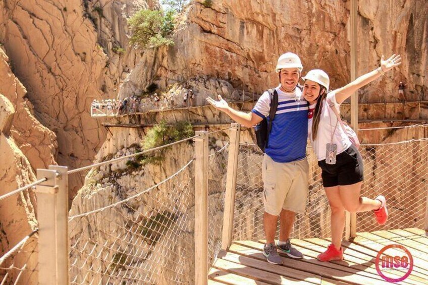 A couple on a wooden bridge with metal railings, surrounded by rocky cliffs. 2