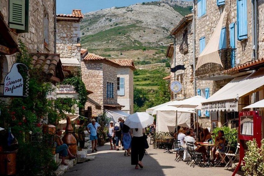Una calle estrecha en un pueblo con edificios de piedra, cafés al aire libre y gente caminando. 5