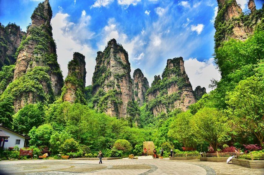 A park with tall rock formations, lush greenery, and a paved area with people walking. 4