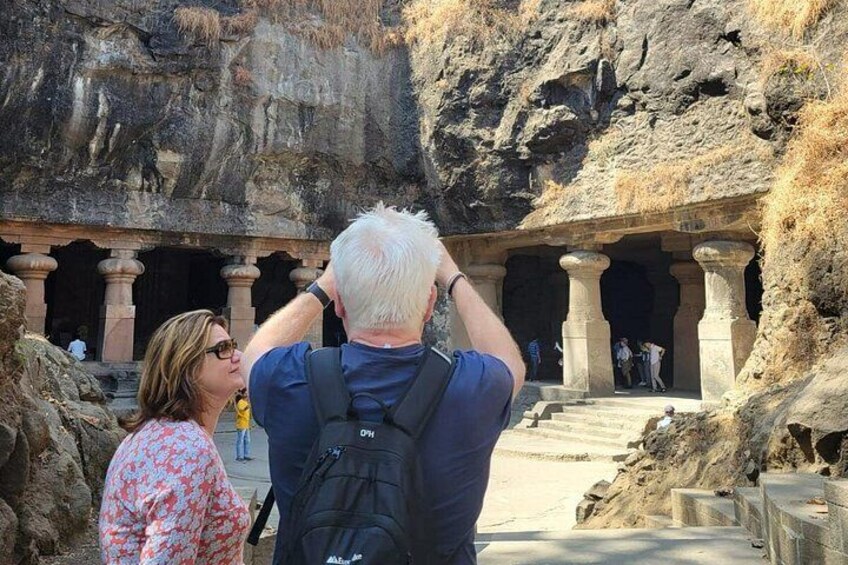 A man taking a photo of ancient rock-cut architecture with columns and a cave entrance. 5