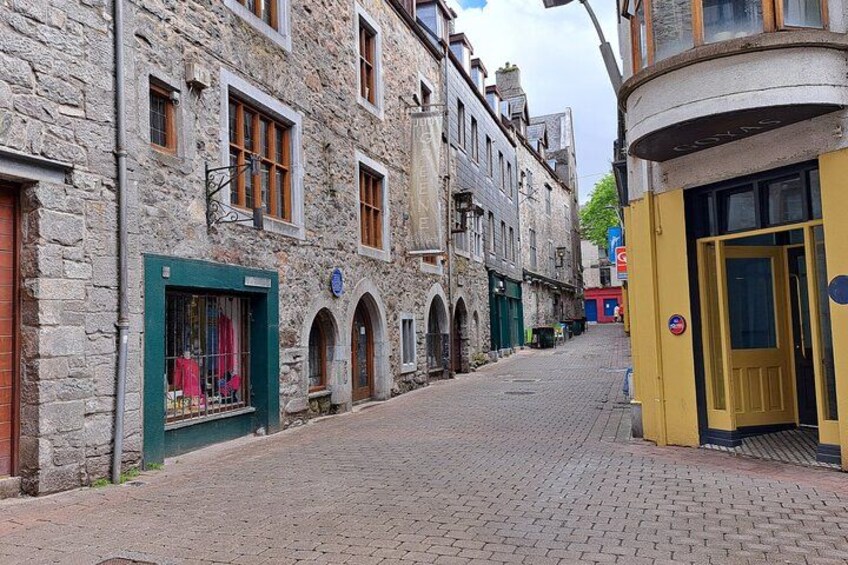 Une rue pavée bordée de bâtiments historiques en pierre, dont certains sont dotés de fenêtres en arc de cercle et de devantures de magasins. 3