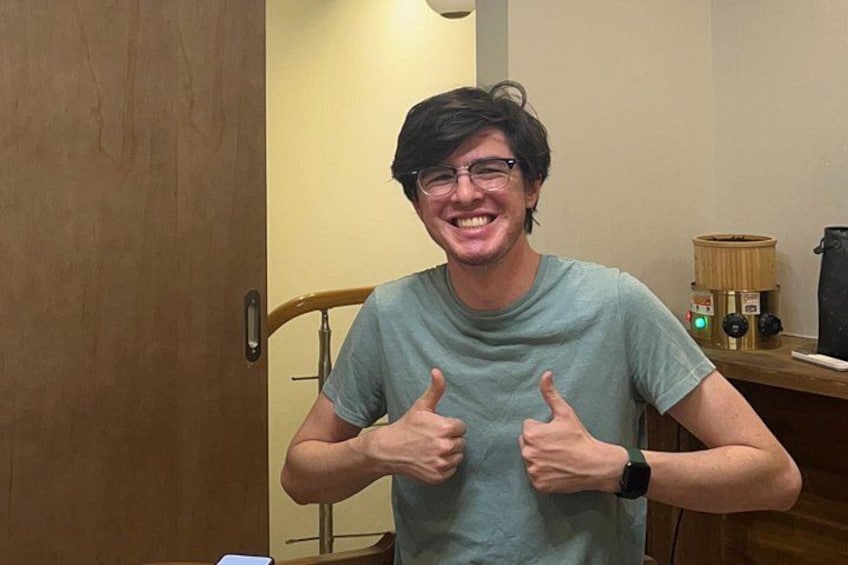 A person giving a thumbs-up gesture in a room with a wooden chair, a wooden door, and a small table with a lamp and a black object on it. 3