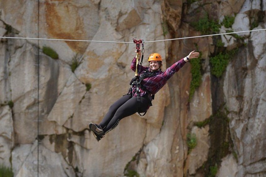 Una persona che vola su una zip-line, allacciata con imbracatura e casco, sospesa a mezz'aria con una scogliera rocciosa sullo sfondo. 2