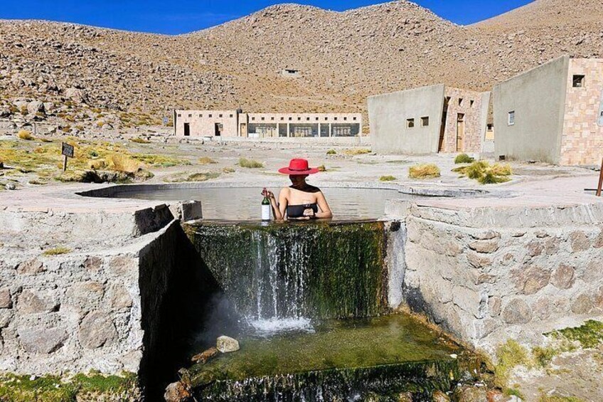 A person in a red hat sitting by a small waterfall in a dry, rocky landscape with buildings in the background. 3