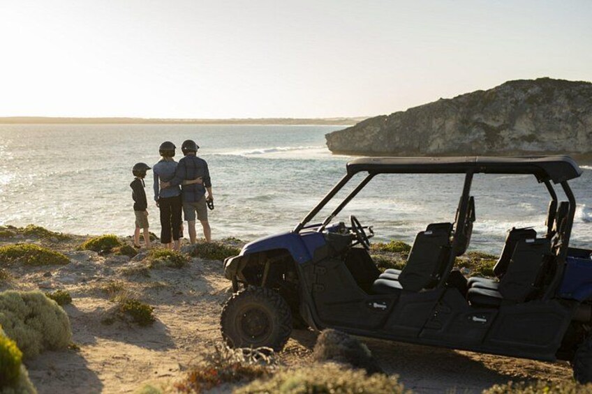 A beach with a dune buggy parked on the sand, a rocky outcrop in the background, and a family standing by the water. 2