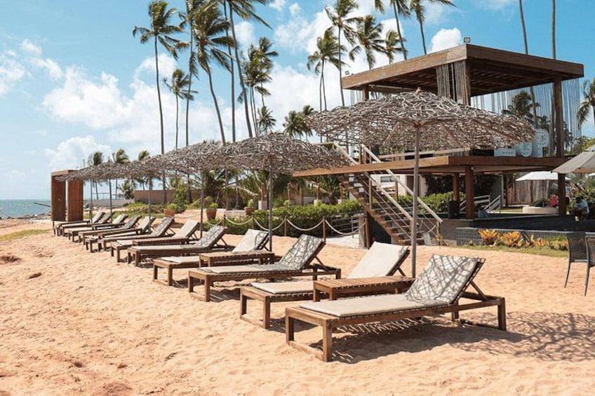 A beach with sun loungers, palm trees, and a wooden structure with umbrellas. 5