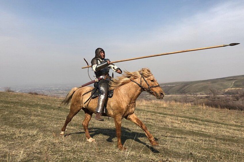Ein in Ritterrüstung gekleideter Reiter auf einem Pferd in einem grasbewachsen Feld, im Hintergrund sanfte Hügel. 4