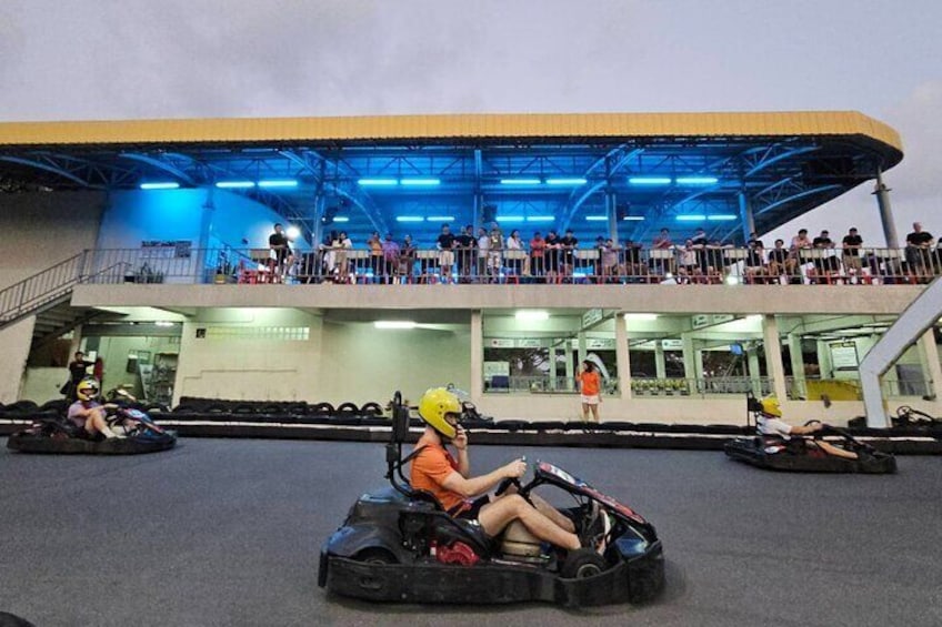 Des personnes conduisent des karts sur une piste, tandis que des spectateurs les observent depuis un balcon. 1