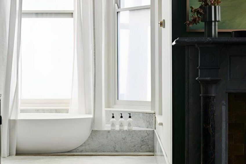 A bathroom with a white bathtub, a window with white curtains, and a black fireplace. 2