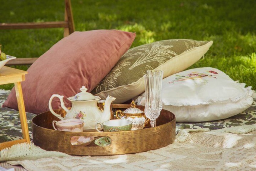 A picnic setup with a teapot, cups, a vase, and decorative pillows on a blanket. 5