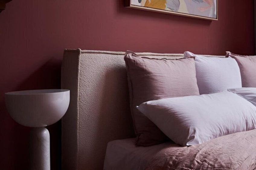 A bed with pink bedding and pillows, a white bedside table, and a wall-mounted painting. 1