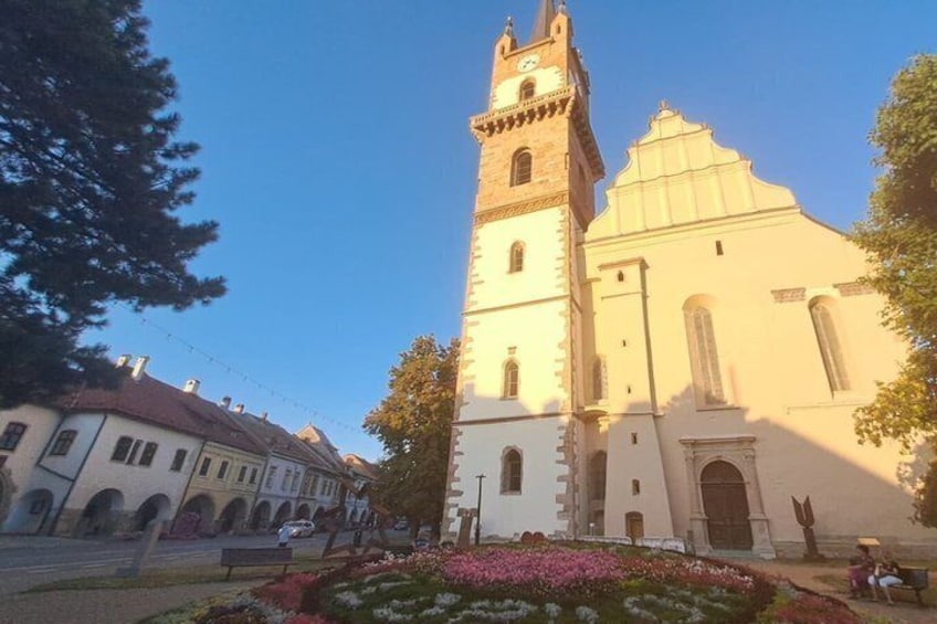 A church with a clock tower, surrounded by buildings and a flower bed. 1