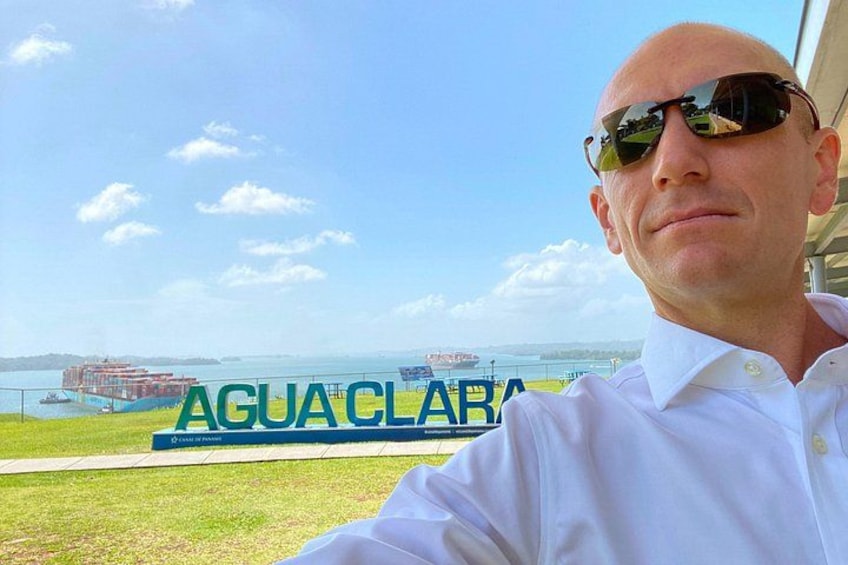 A man in a white shirt standing in front of a sign that reads 'AGUACLARA' with a body of water and boats in the background. 1