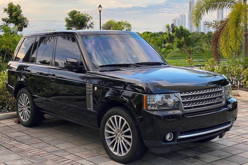 A black SUV parked on a paved area with greenery and buildings in the background. 3