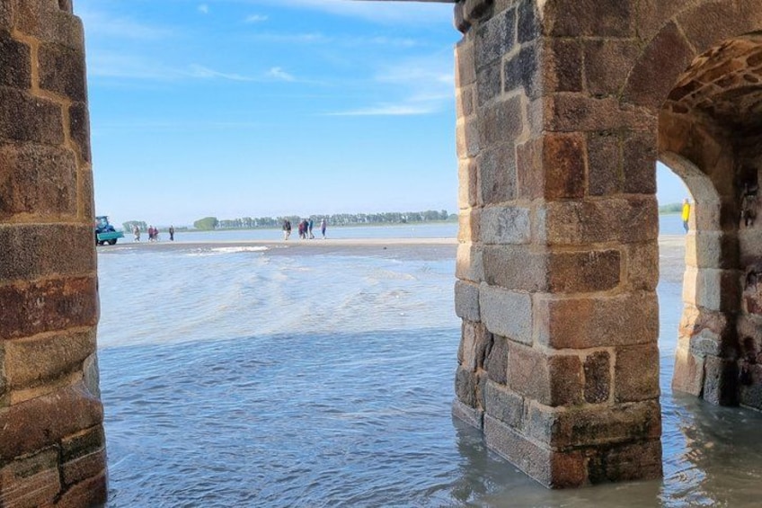 A stone bridge over water with people walking on the beach in the background. 2