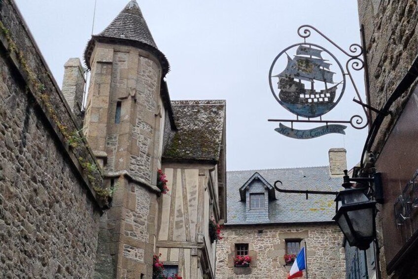 A narrow street with stone buildings, a tower, and a sign with a ship. 2