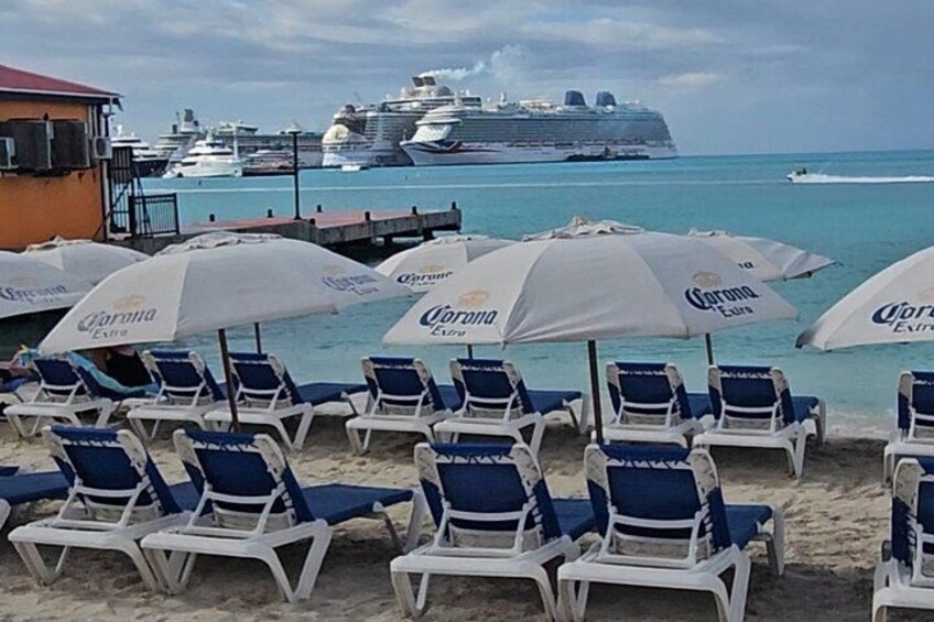 Beach chairs and umbrellas with the Corona logo, a cruise ship, and boats in the background. 1