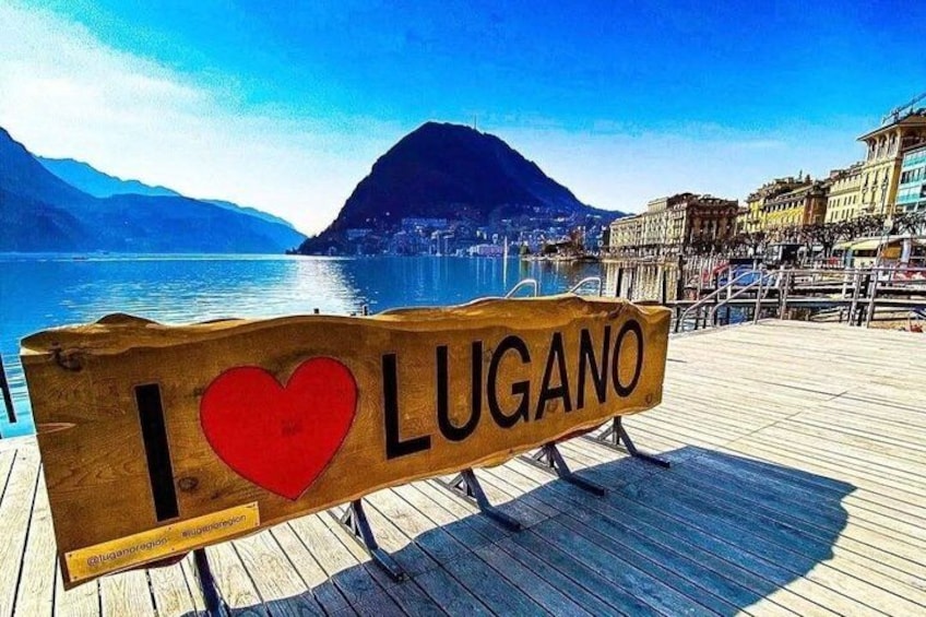 Un letrero de madera con 'I ♥ LUGANO' escrito, con un paisaje pintoresco de un lago y montañas al fondo. 5