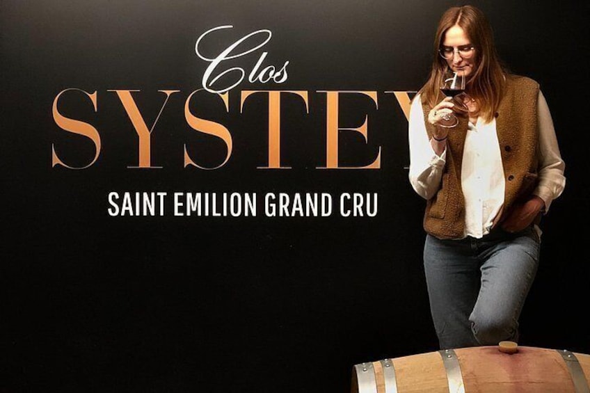 Eine Person steht neben einem Holzfass, hält ein Weinglas und im Hintergrund ist Text zu sehen: 'Clos SYSTEY SAINT EMILION GRAND CRU'. 4