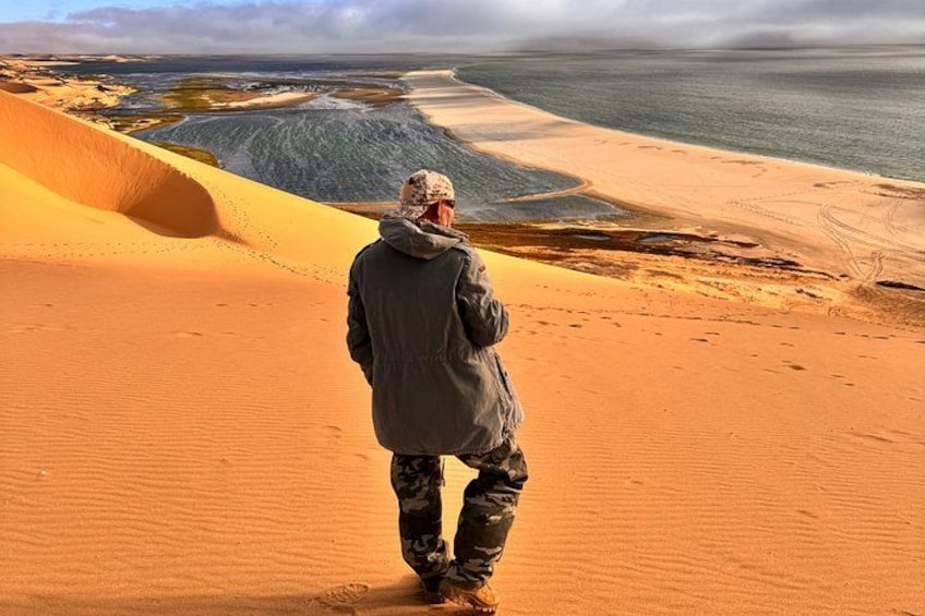 Eine Person steht auf einer Düne und blickt auf eine Küstenlandschaft mit Dünen und einem Gewässer. 1