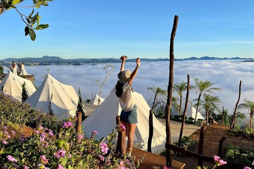Eine Person steht auf einer Holzdielen Terrasse mit Blick auf weiße Zelte und Palmen, die über ein Tal blicken. 1