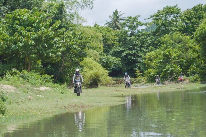 Motorcyclists riding along a riverbank surrounded by lush greenery. 5