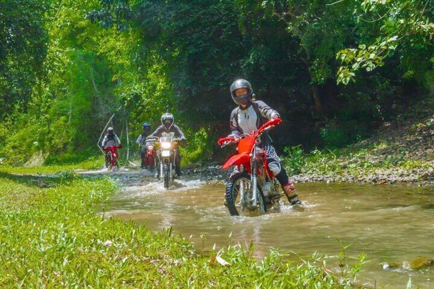 Three motorcyclists riding through a shallow water crossing in a forested area. 1