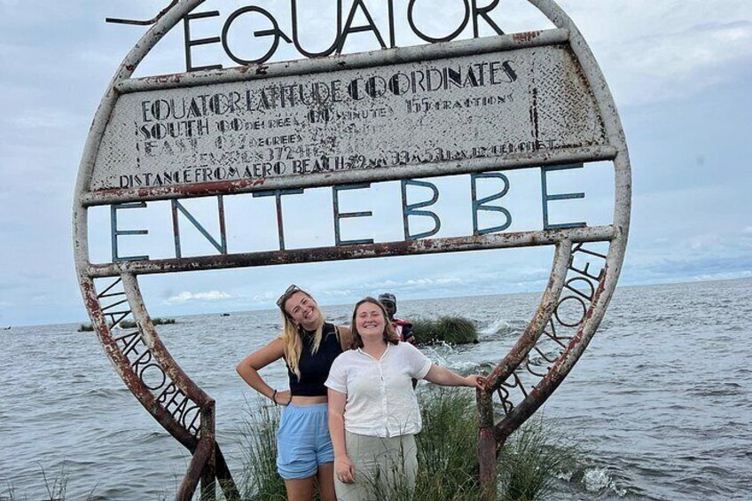 Un letrero con los textos 'EQUATOR' y 'ENTERBEE', dos personas de pie frente a él, y un cuerpo de agua al fondo. 5