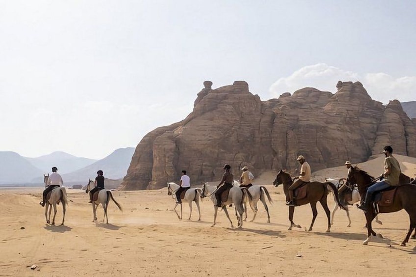 A group of people riding horses in a desert landscape with large rock formations in the background. 2