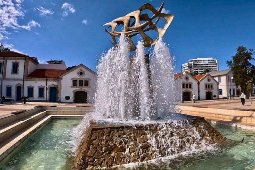 Una fuente con chorros de agua, una gran escultura abstracta y edificios históricos al fondo. 1