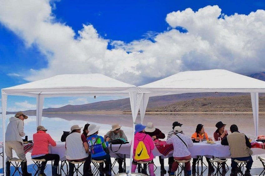 A group of people sitting under white tents at a lakeside with mountains in the background. 3
