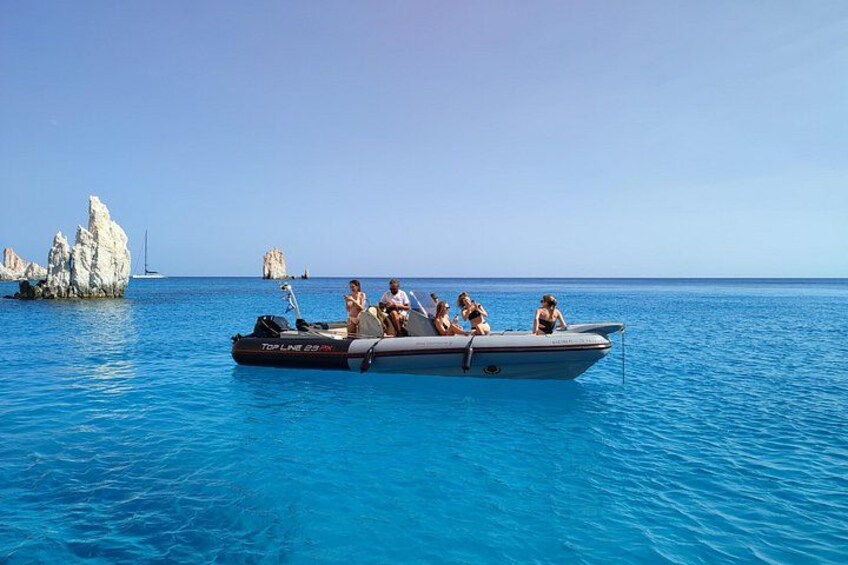 Een boot met mensen op een heldere blauwe zee, gesteente en een zeilboot in de verte. 5