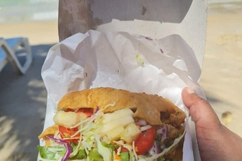 En hånd holder en sandwich med salat, tomat og en friturestegt ret, sandsynligvis kylling, på en bolle, med en strand i baggrunden. 2