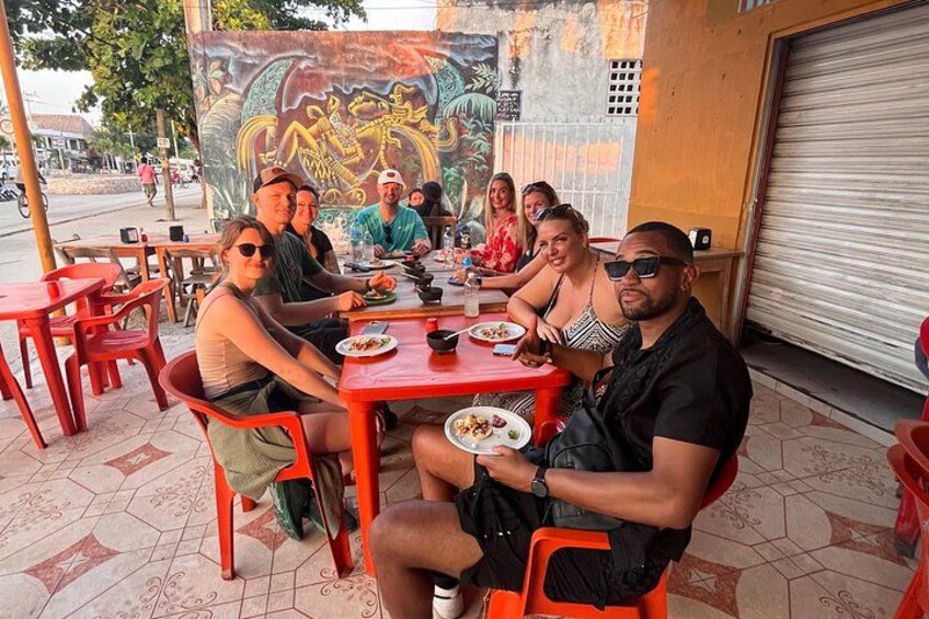 A group of people dining at an outdoor restaurant with red chairs and tables, a mural in the background, and a street visible in the distance. 2
