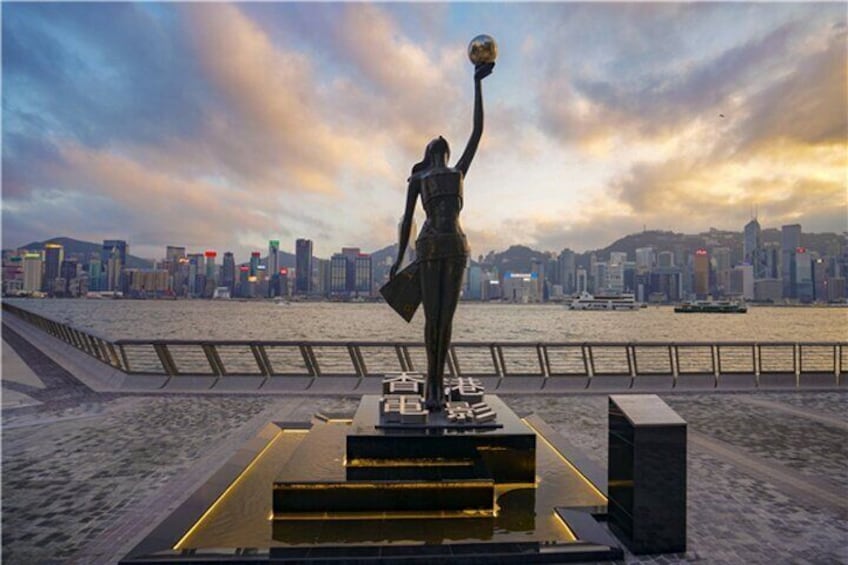 En statue af en kvinde der holder en globe på en platform med trapper, med udsigt over en havnefront og en bysilhuet i baggrunden. 1