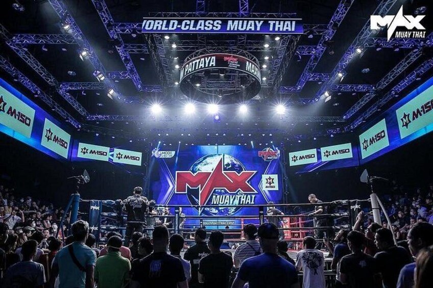 En boksring med et publikum, et stort skærmbillede der viser 'MAX Muay Thai' og 'PATTAYA', samt branding for arrangementet. 3