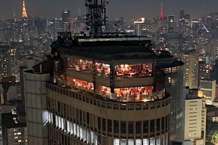 Un edificio de varios pisos con interiores iluminados, rodeado por un paisaje urbano con otros edificios y una torre distante. 2