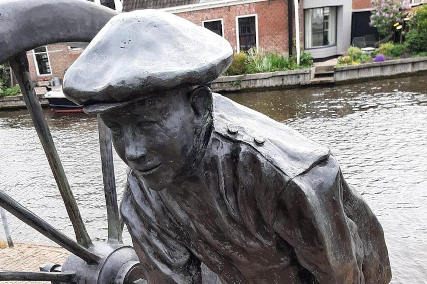 A bronze statue of a man wearing a hat and a coat, positioned near a canal with a boat in the background. 5